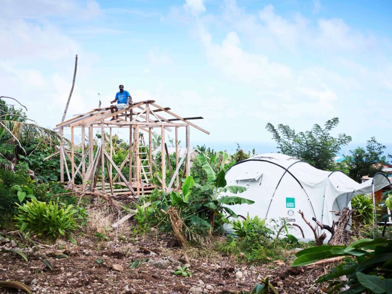 Man sitting on top of a part built wooden house frame beside a shelter box tent