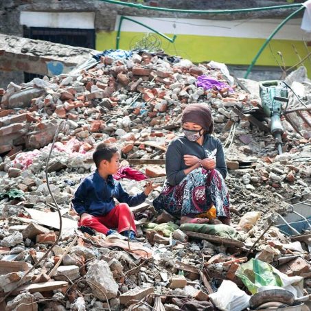 Eine Frau mit Maske sitzt mit einem kleinen Jungen auf Trümmern und Schutt in Nepal.