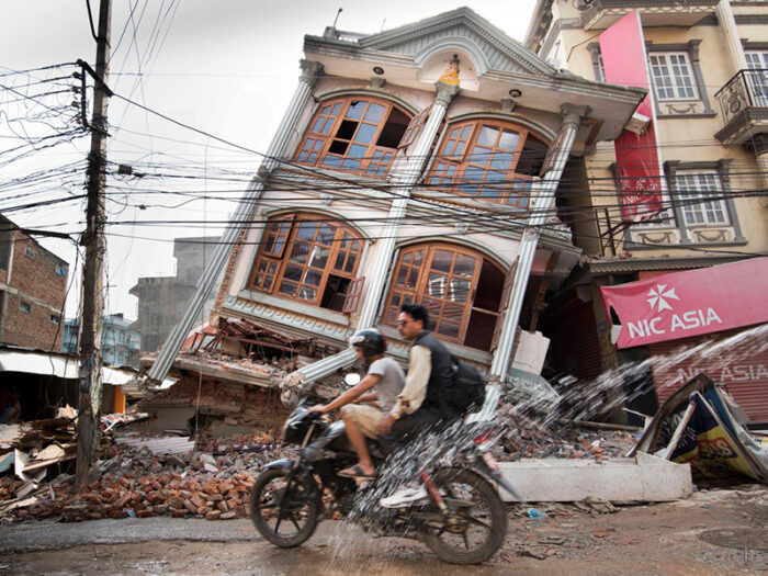 2 people on a motorcycle pass by a tilted, destroyed building in Nepal after the 2015 earthquake
