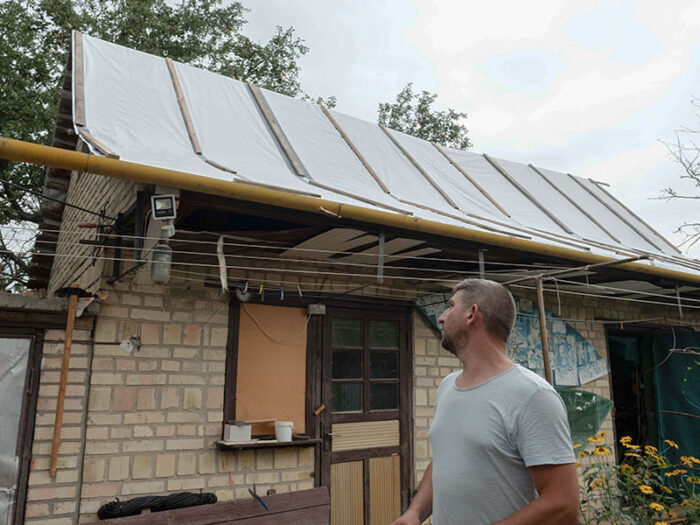 Ein ukrainischer Mann in grauem T-Shirt blickt auf sein Haus. Das Dach wurde nach Bombardierungen mit Planen repariert