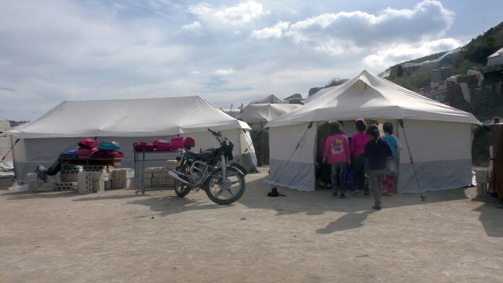 Zwei große weiße Zelte stehen auf sandigem Boden, mit Kindern und einem Motorrad drum herum.