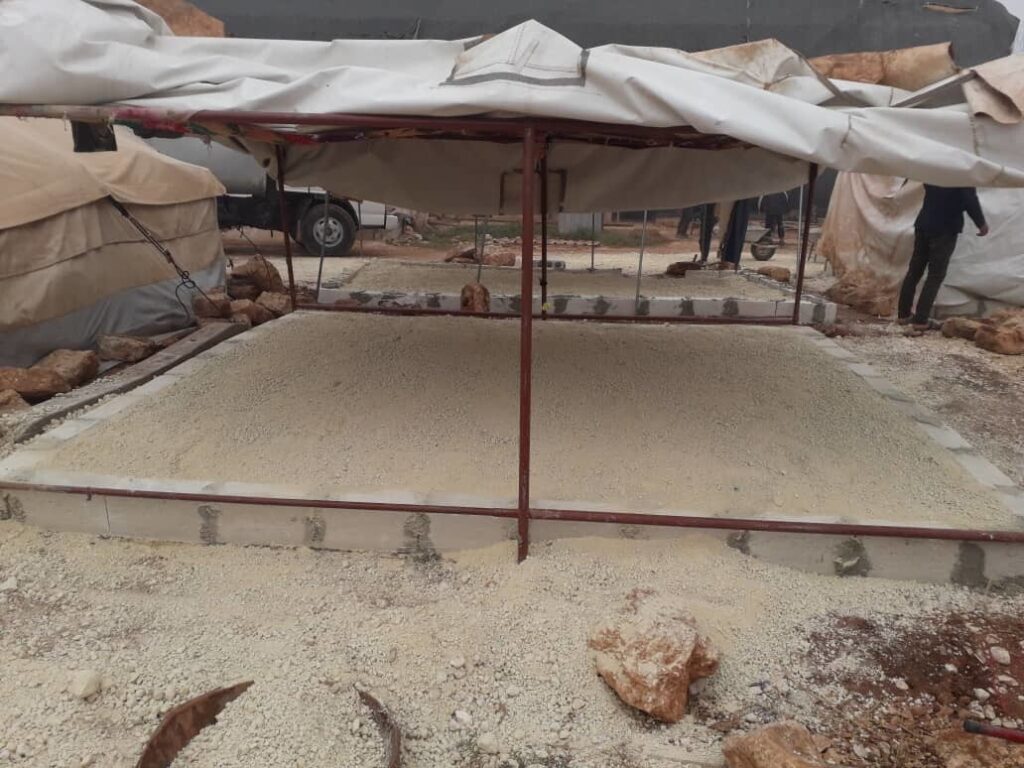 Ein Quadrat aus Ziegelsteinen, das mit Beton gefüllt ist, um eine ebene Fläche zu schaffen, mit darüber an Pfosten aufgehängten Planen.