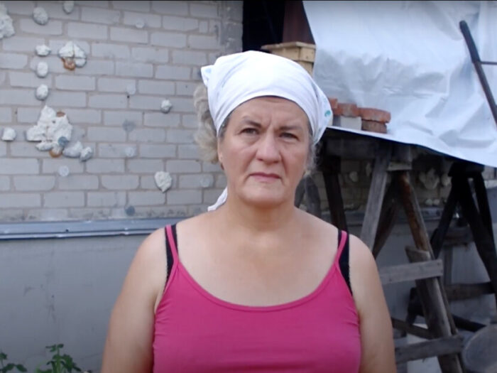 Eine Frau steht vor einem beschädigten Haus in der Ukraine. Ein Teil des Hauses wird mit einer Plane bedeckt.