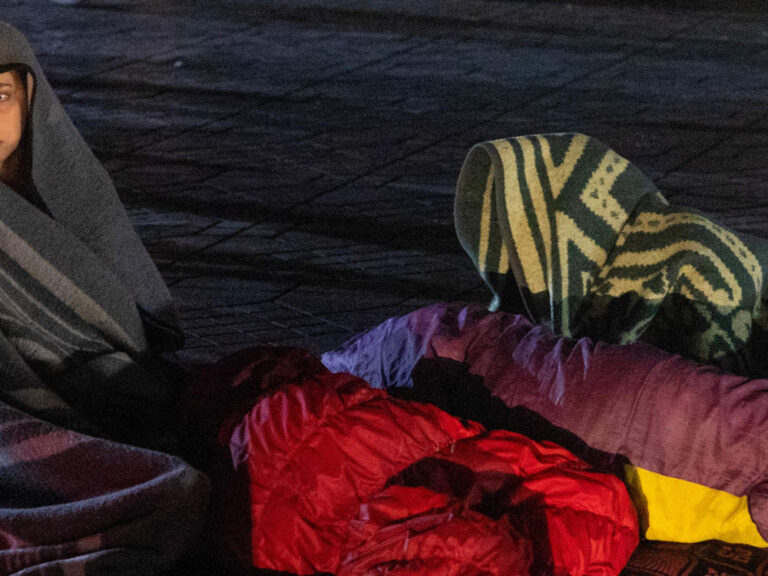 Eine Frau sitzt in eine Decke gewickelt auf dem Boden mit Personen in Schlafsäcken um sich