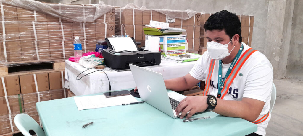 Mann mit Gesichtsmaske, der mit einem Laptop an einem Schreibtisch im Lager von ShelterBox Operations Philippines sitzt.