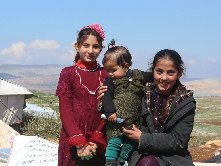 Drei Kinder in Winterkleidung stehen vor einem Zelt in der syrischen Landschaft