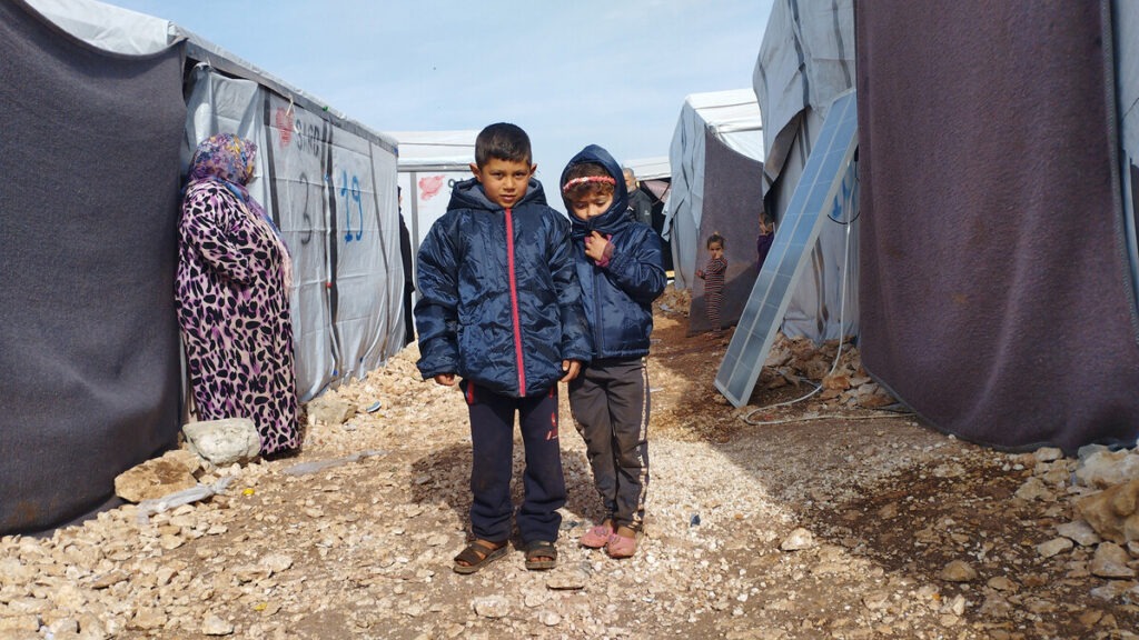Zwei junge Kinder in Winterjacken stehen inmitten von Unterkünften in Syrien