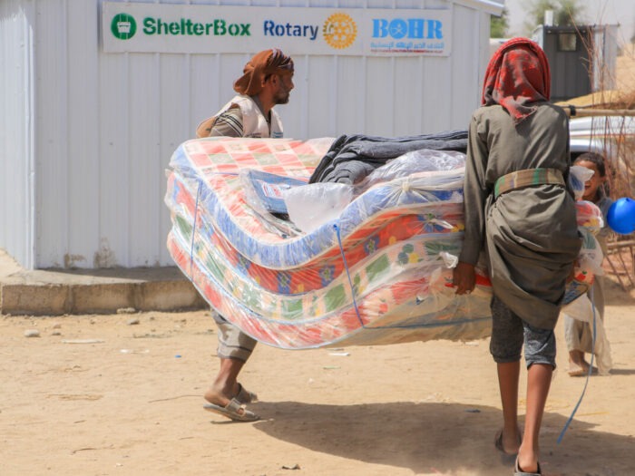 People carrying mattresses next to an iron net shelter with the ShelterBox logo in Yemen