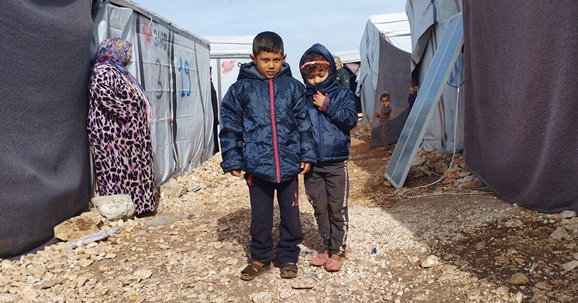 Zwei junge Kinder stehen inmitten eines Flüchtlingslagers und tragen Wintermäntel.
