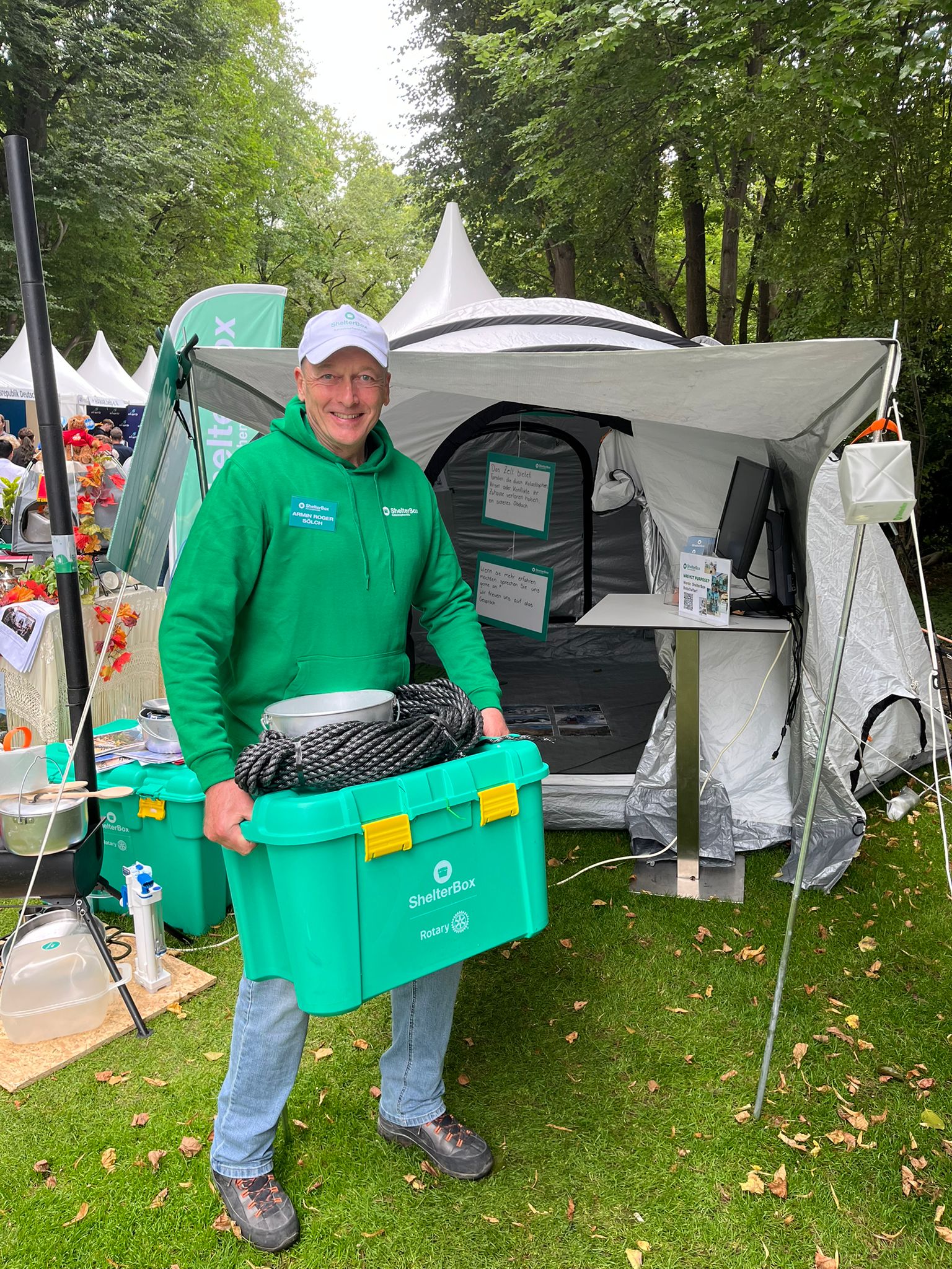 Ein Mann in grünem Kapuzenpulli steht vor einem Zelt und hält eine grüne ShelterBox Kiste in den Händen